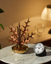 Decorative diffuser with organic wooden leaves and a polished gold base by Alessi, on a white marble table next to a black alarm clock.