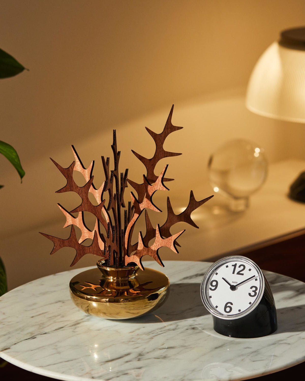 Decorative diffuser with organic wooden leaves and a polished gold base by Alessi, on a white marble table next to a black alarm clock.