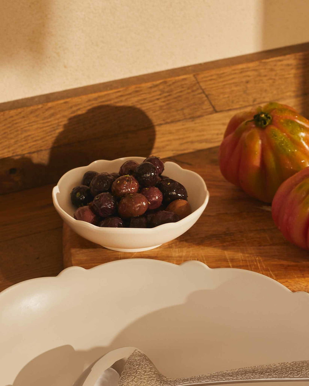 Scalloped white serving bowl by Alessi, filled with dark olives, next to heirloom tomatoes on a wooden board.