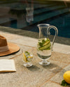 Clear glass pitcher by Alessi with cucumber, lemon, and mint infused water, next to a matching glass, book, and hat by a pool.