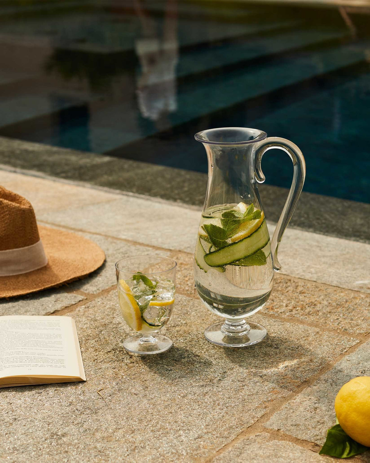 Clear glass pitcher by Alessi with cucumber, lemon, and mint infused water, next to a matching glass, book, and hat by a pool.
