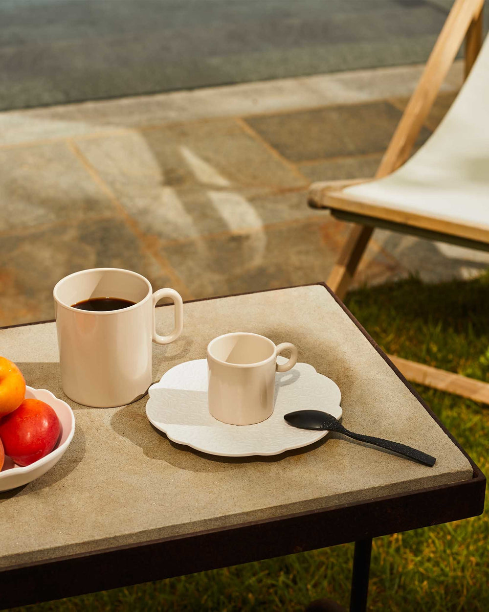 White scalloped plate with relief pattern by Alessi and light beige mugs on a textured outdoor table.