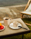 Beige ceramic mugs and patterned saucer by Alessi on an outdoor stone table with fresh fruit and a deck chair.
