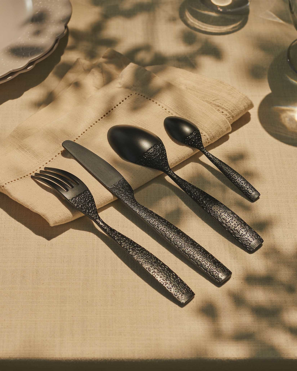 Black metal cutlery set by Alessi with ornate textured handles, arranged on a beige napkin.