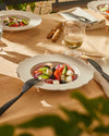 Off-white scalloped plates and black textured cutlery by Alessi, holding a fresh salad on an outdoor table.