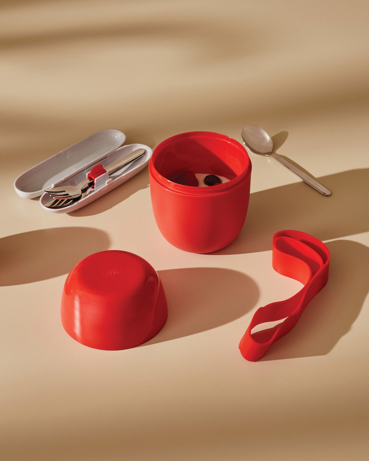 Red Food à porter portable food container by Alessi, open with its lid, a cutlery case, and a silicone strap.