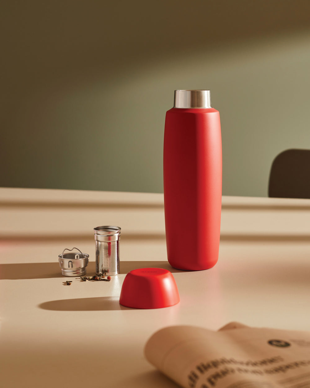Red thermos bottle by Alessi, standing upright on a beige table next to its red lid, a metal tea infuser, and loose tea leaves.