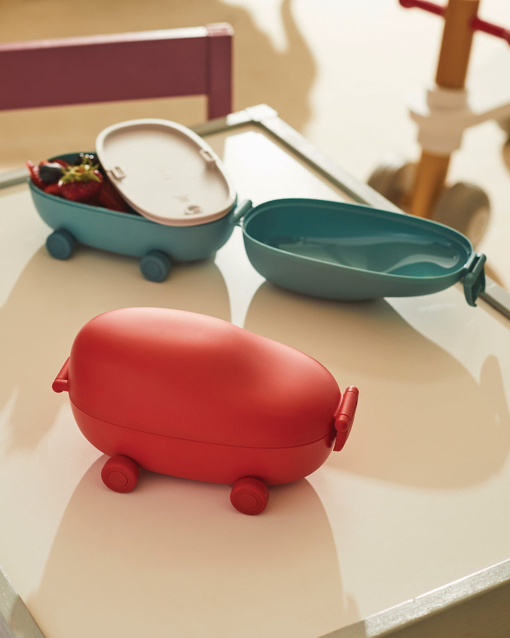Red oval lunchbox by Alessi with wheels, alongside two open teal food containers, one with fruit.