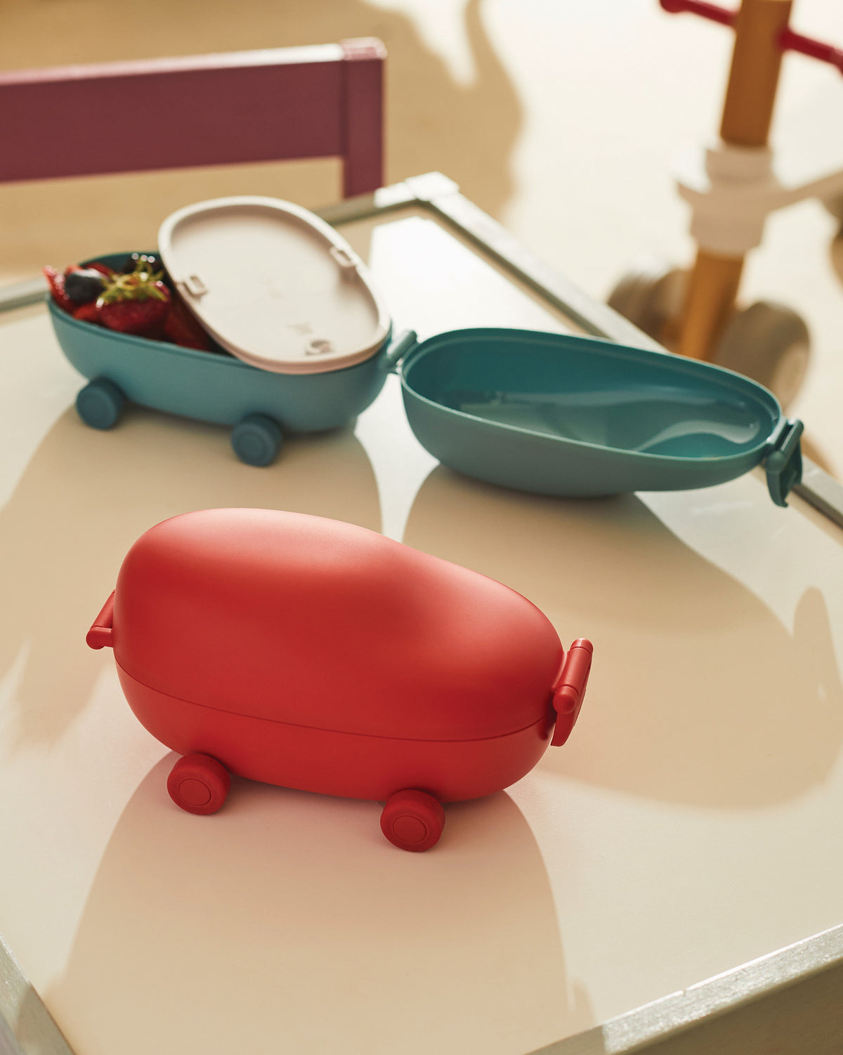 Red oval lunchbox by Alessi with wheels, alongside two open teal food containers, one with fruit.