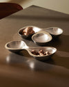 Silver-toned double-section serving bowls by Alessi, with a matte exterior and shiny interior, holding walnuts and hazelnuts.