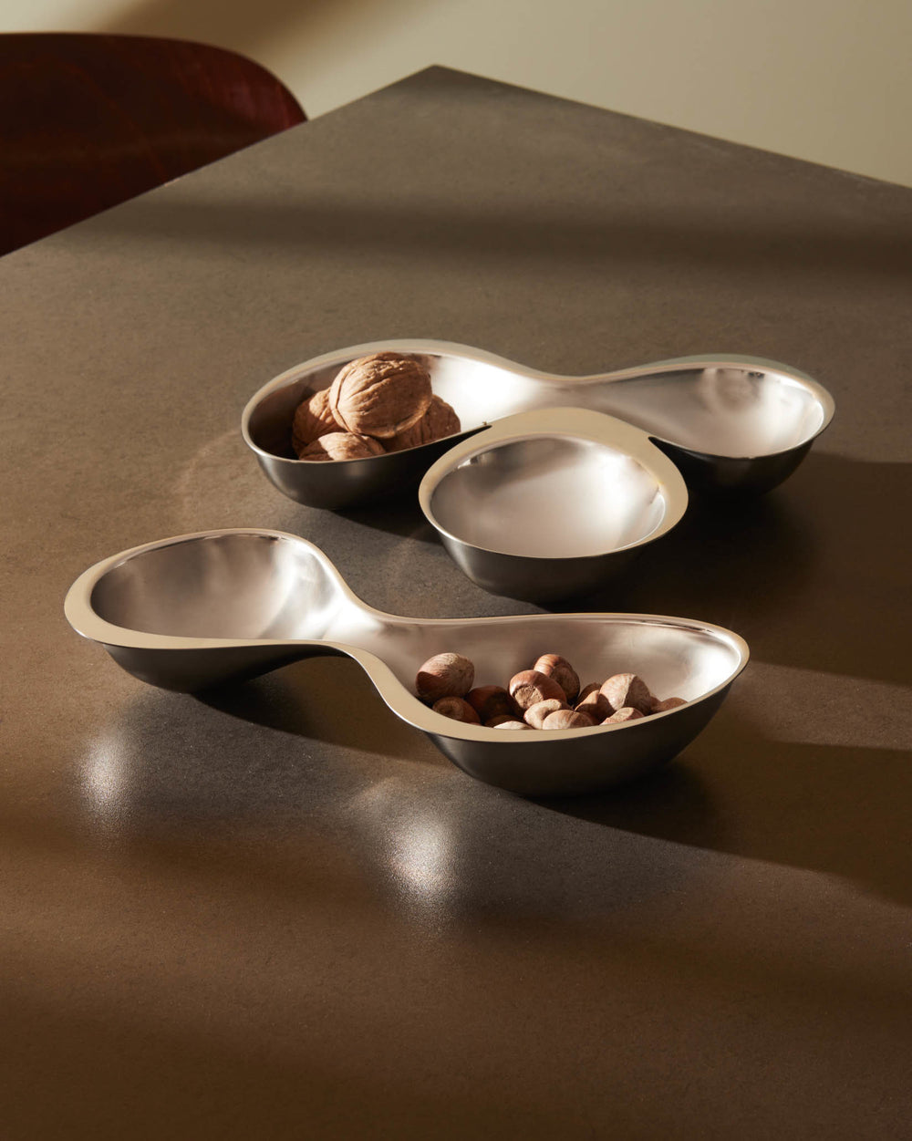 Silver-toned double-section serving bowls by Alessi, with a matte exterior and shiny interior, holding walnuts and hazelnuts.