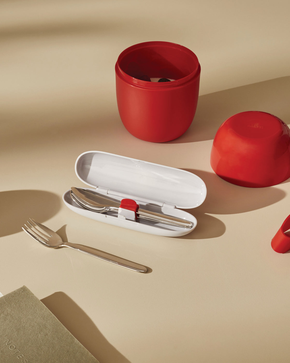 Red portable food carrier and silver cutlery set by Alessi, with a white cutlery case and a notebook on a light tan surface.