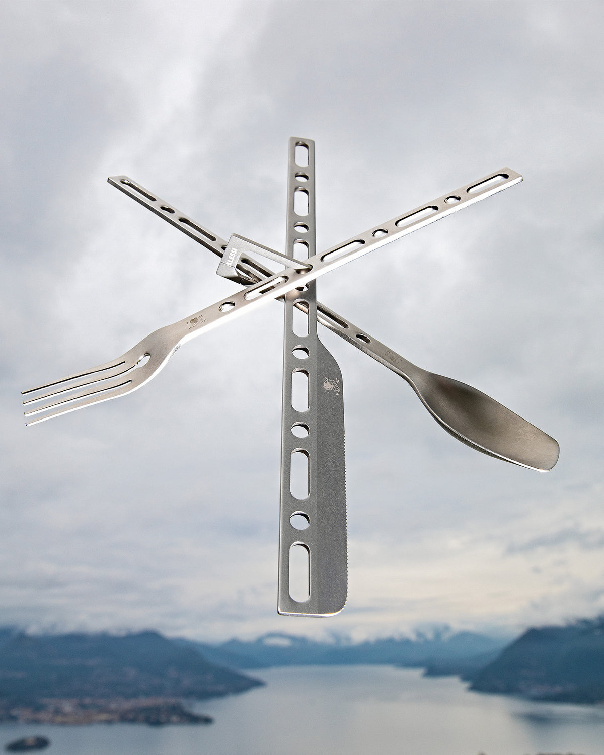 A grey metal modular travel cutlery set by Alessi, with a fork, serrated knife, and spoon arranged in a cross, against a blurred mountain lake.