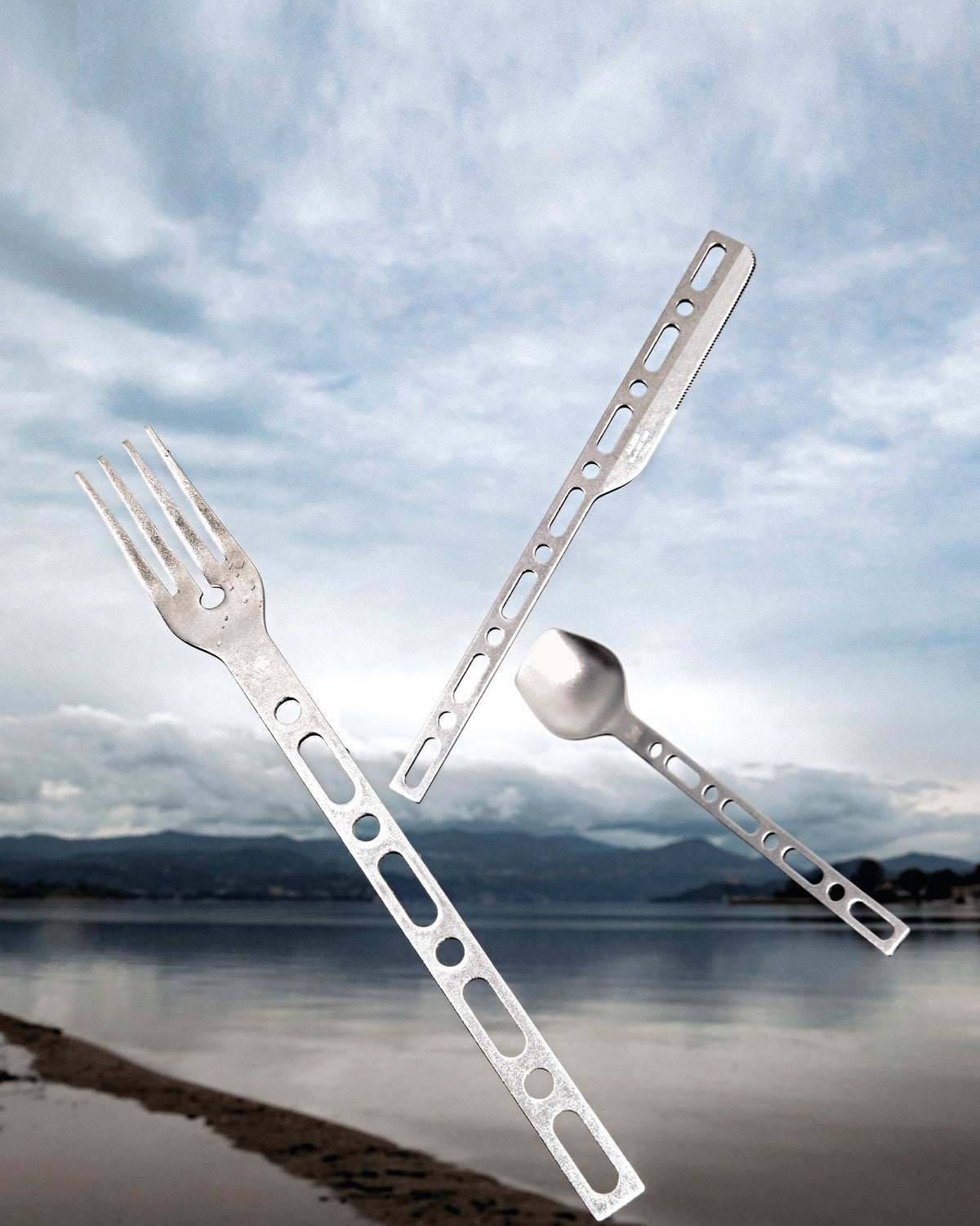 Silver metal camping utensil set by Alessi, featuring a fork, knife, and spoon with cut-out handles, floating against a lake and cloudy sky.