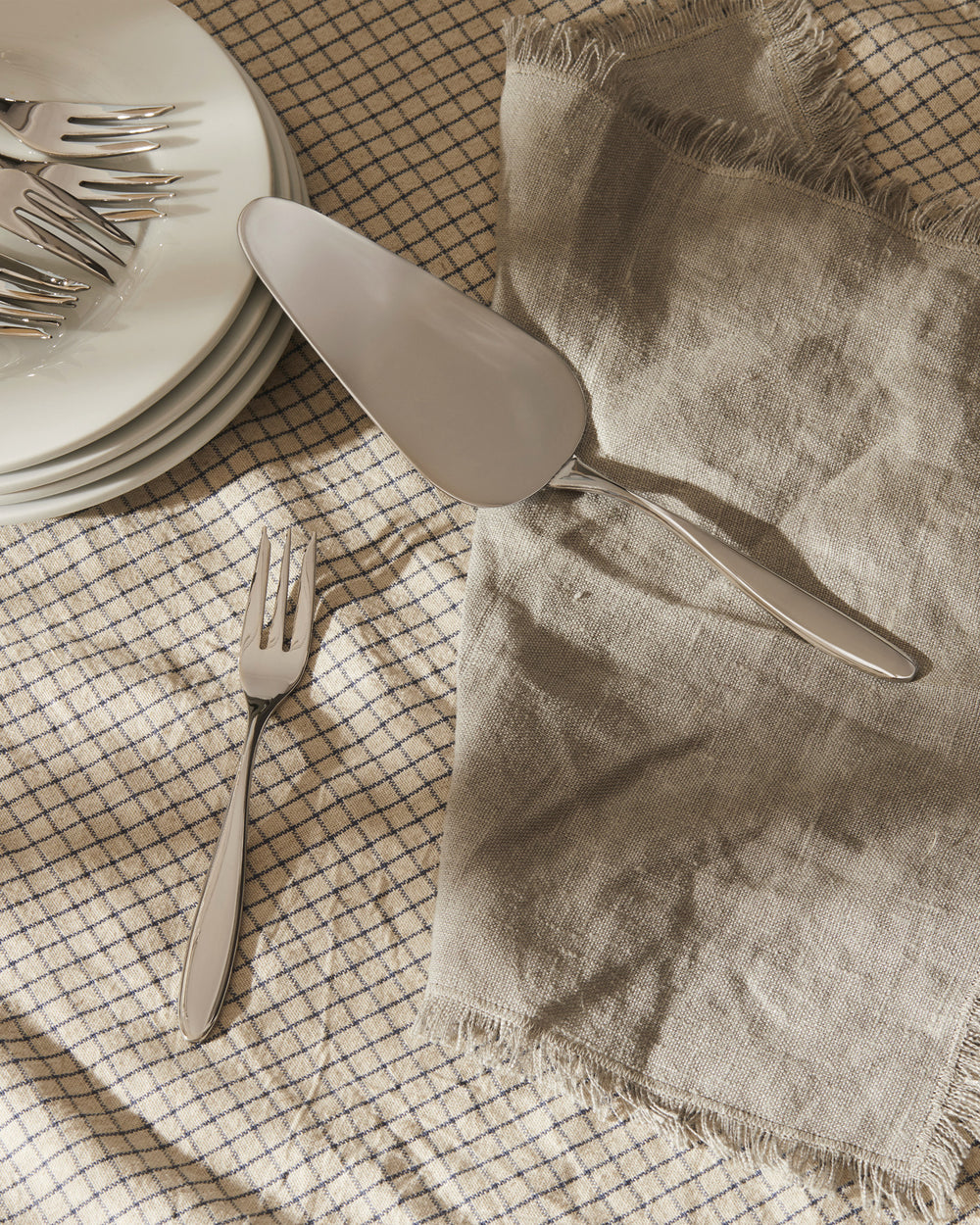 Shiny silver Mami cake server by Alessi, with a matching fork, stacked plates, and a linen napkin on a plaid tablecloth.