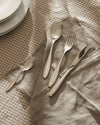 Four-piece polished silver cutlery set by Alessi on a fringed grey linen napkin and plaid tablecloth.