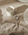 White ceramic mug by Alessi on a plaid tablecloth with a silver spoon and plate.