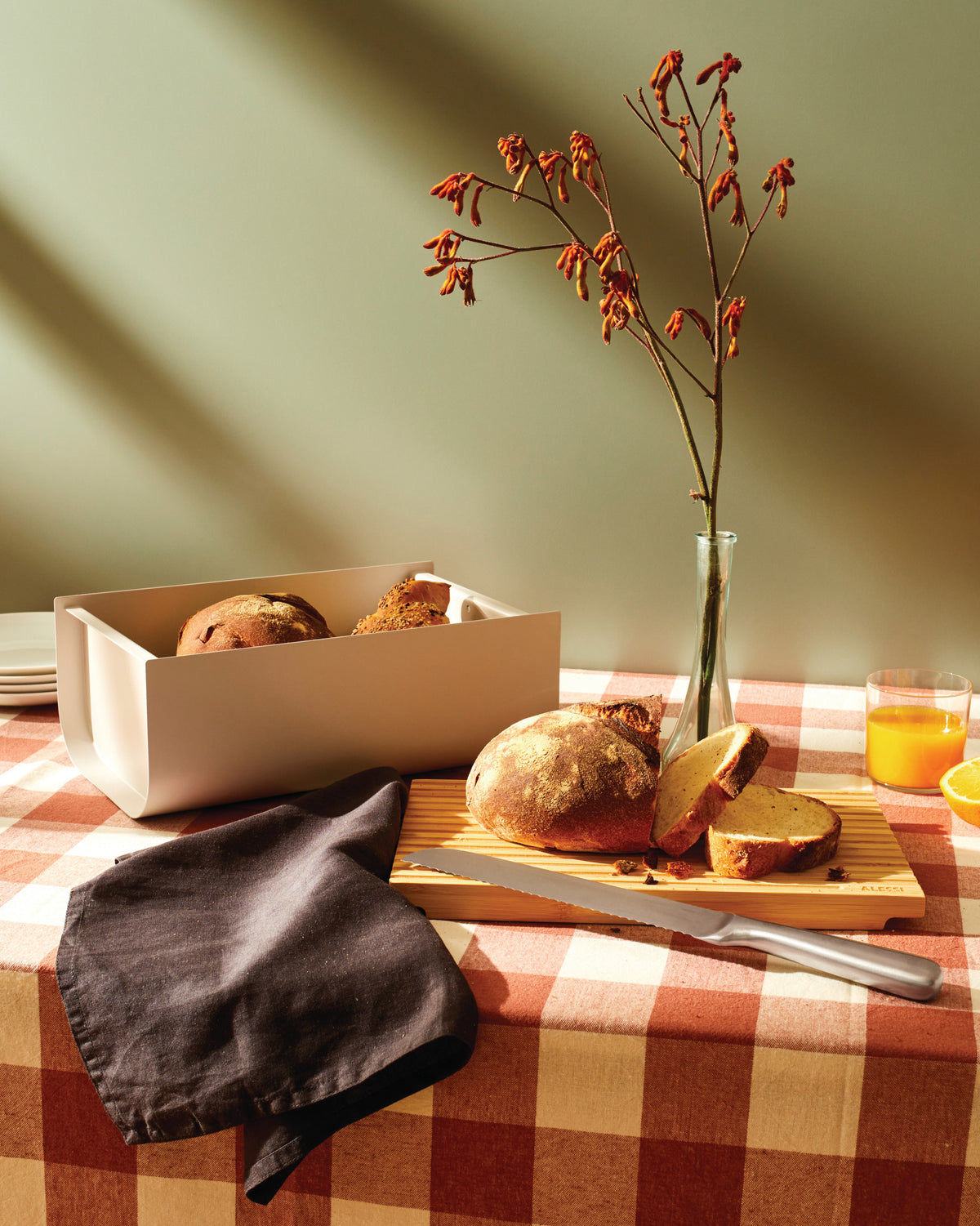 Light beige Mattina bread box by Alessi holding artisan loaves on a red checkered tablecloth.
