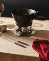Black cast iron fondue set by Alessi, with marked serving and dipping forks on a chrome stand, beside red wine and a red linen napkin.