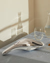 Sleek silver and white handheld duster by Alessi with a red button, on blue linen with crumbs, beside a water glass and carafe.