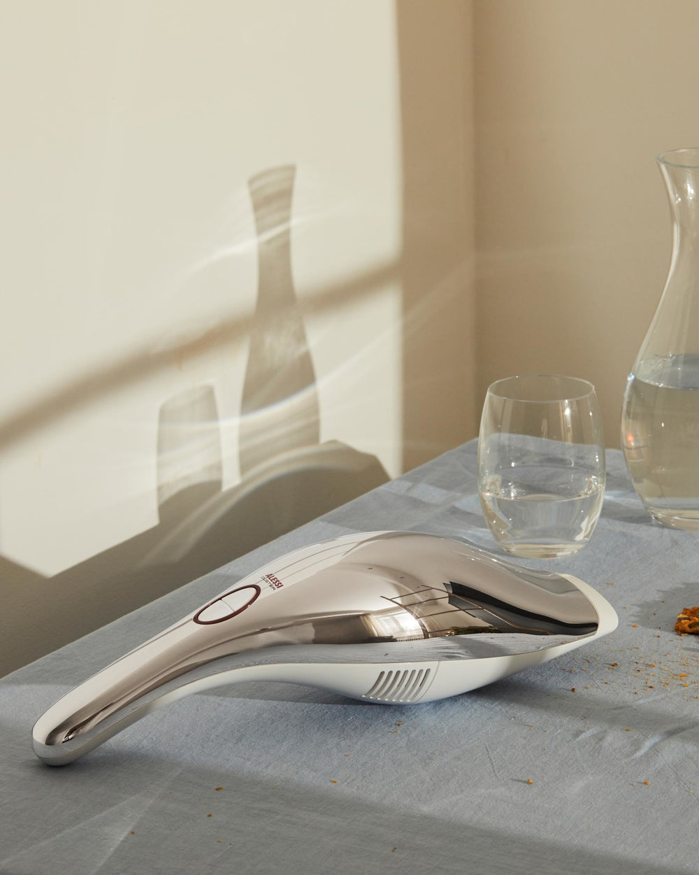 Sleek silver and white handheld duster by Alessi with a red button, on blue linen with crumbs, beside a water glass and carafe.