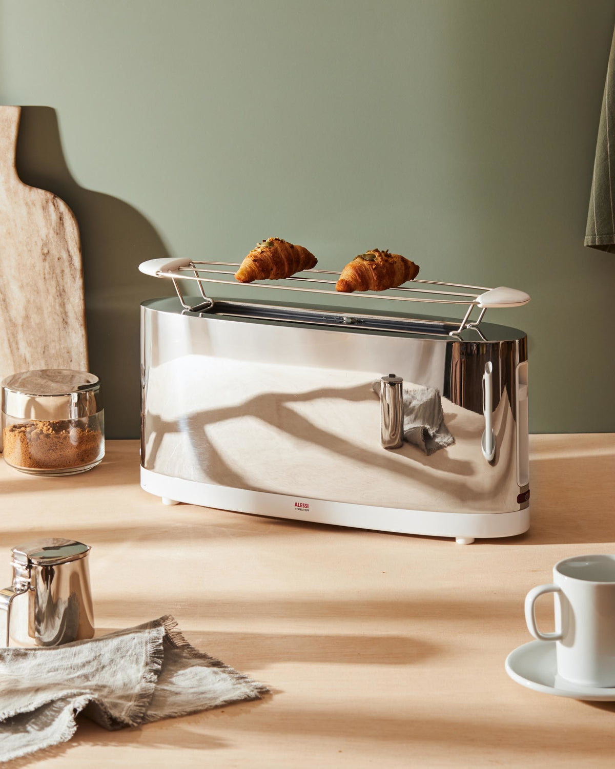 Long silver chrome toaster by Alessi with a white base and two croissants on a warming rack.