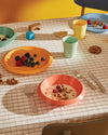 Colorful kids tableware collection by Alessi arranged on a checkered tablecloth with breakfast foods and wooden toys.