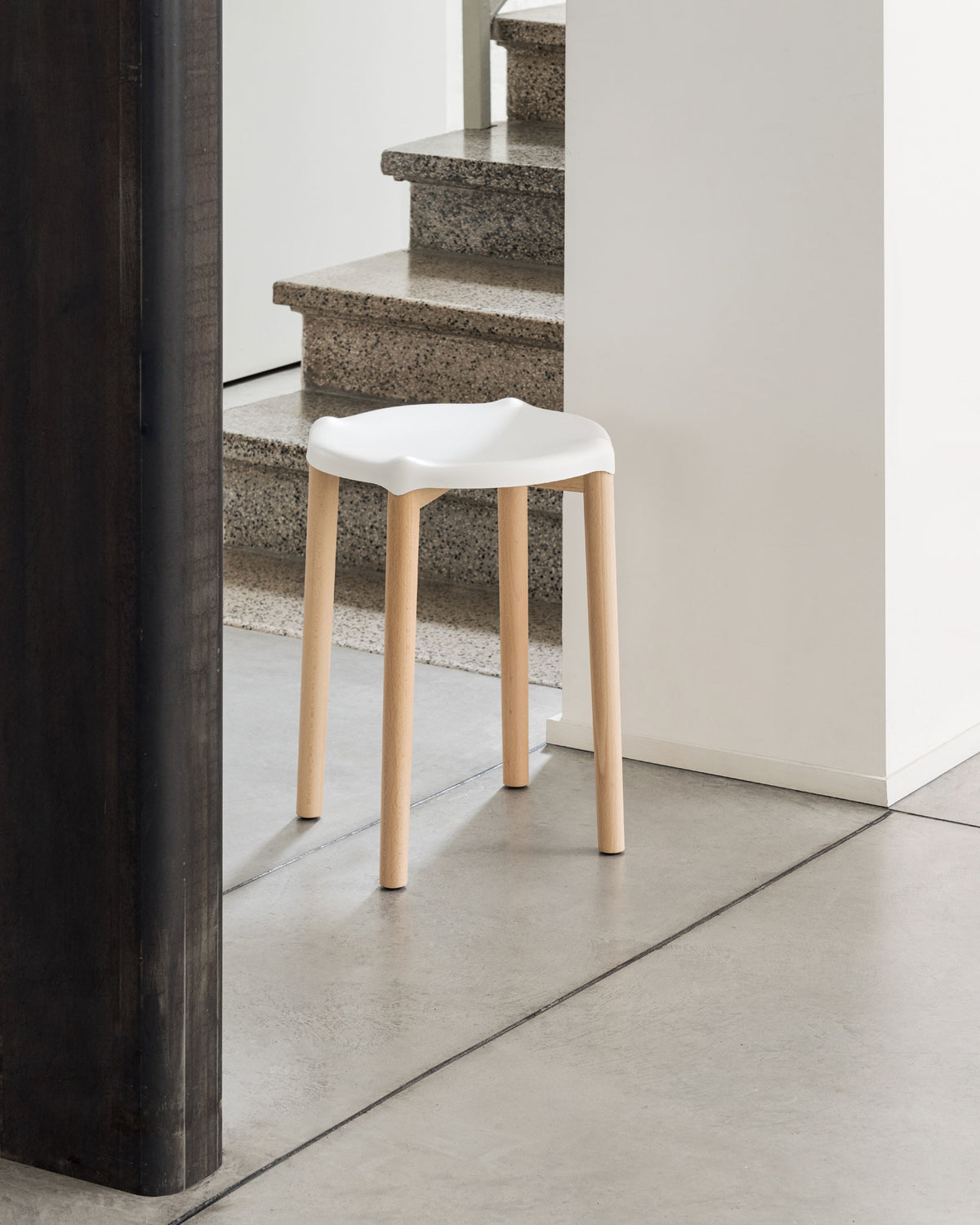 White stool with a contoured seat and four light wood legs by Alessi, on a concrete floor with granite stairs in the background.