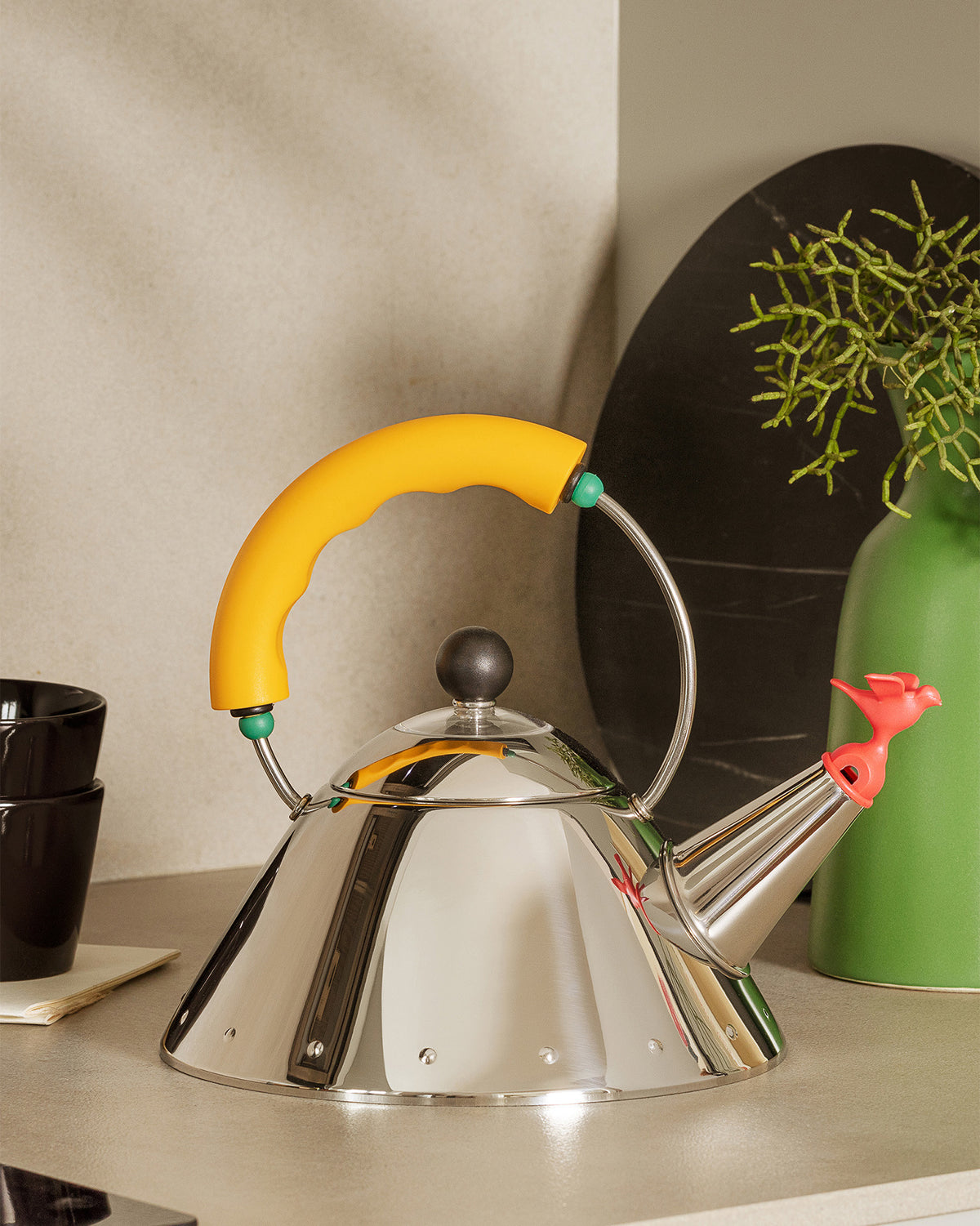 Stainless steel whistling kettle by Alessi with a yellow handle and a red bird-shaped spout.