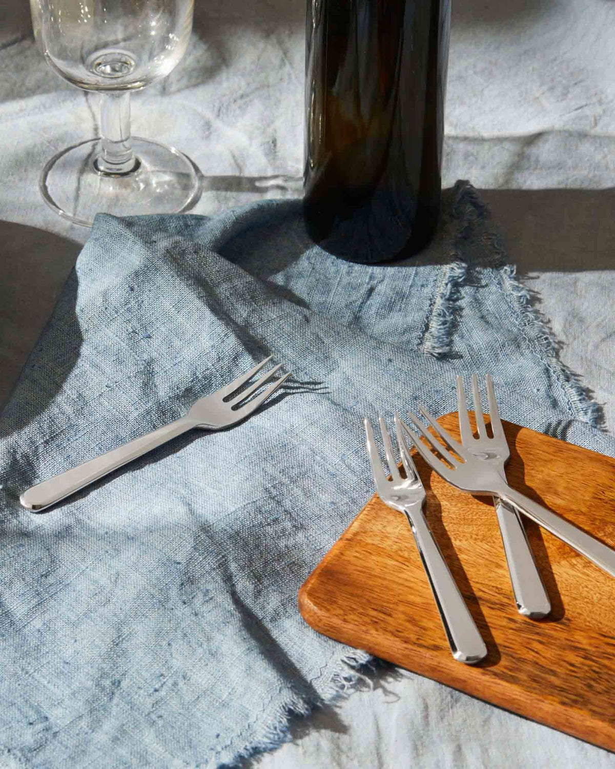 Silver forks by Alessi displayed on a blue linen napkin and wooden board.