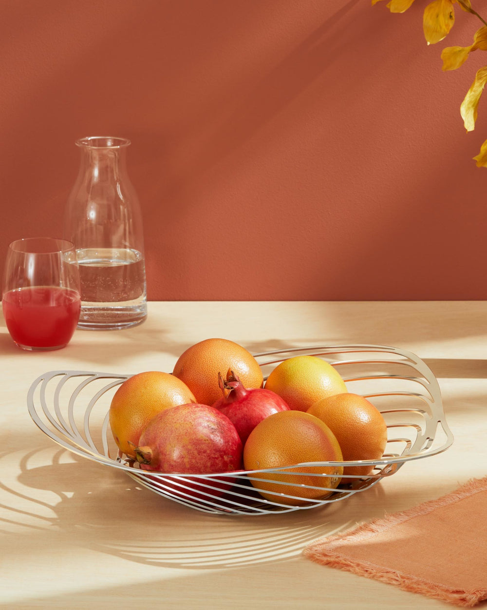 Silver-toned metal Trinity fruit bowl by Alessi with an open, sculptural design, holding grapefruits and pomegranates on a light wooden table.