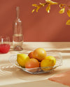 Silver-toned metallic sculptural Trinity fruit bowl by Alessi, holding lemons and grapefruits on a light wood surface.